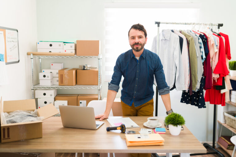 Mannlig nettbutikk-entrepenør står på hjemmelageret sitt med produkter og pakkeesker bak seg.