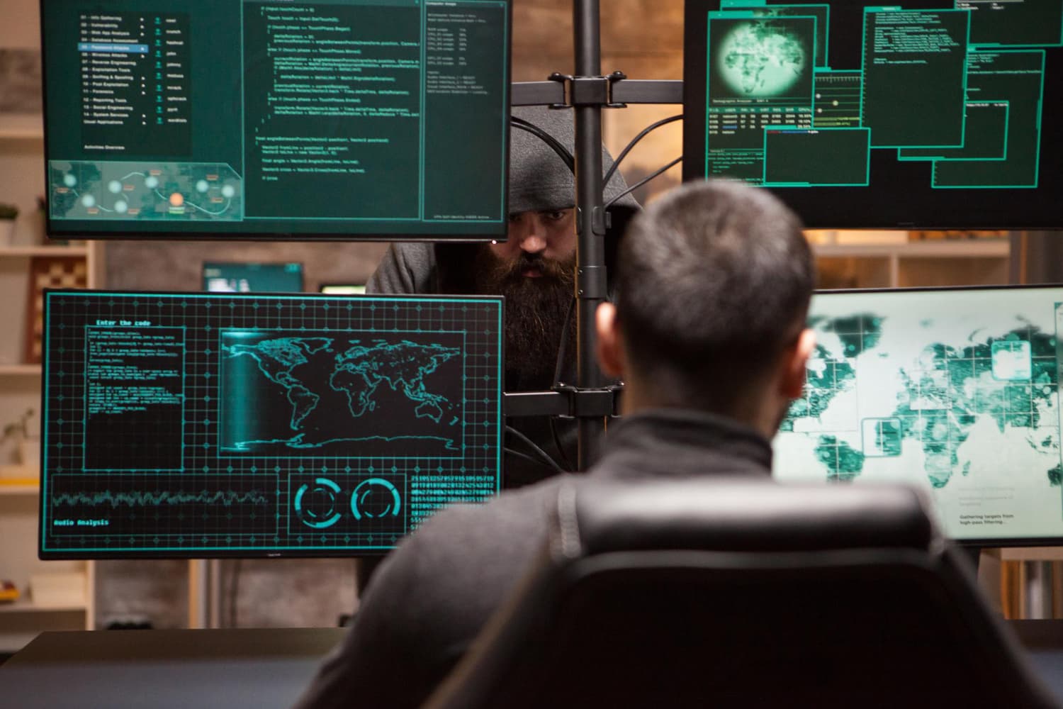 DDOS angrep illustrasjonsbilde - to skumle menn i svarte hettegensere som utfører et hackerangrep med tre skjermer med grønn kode.